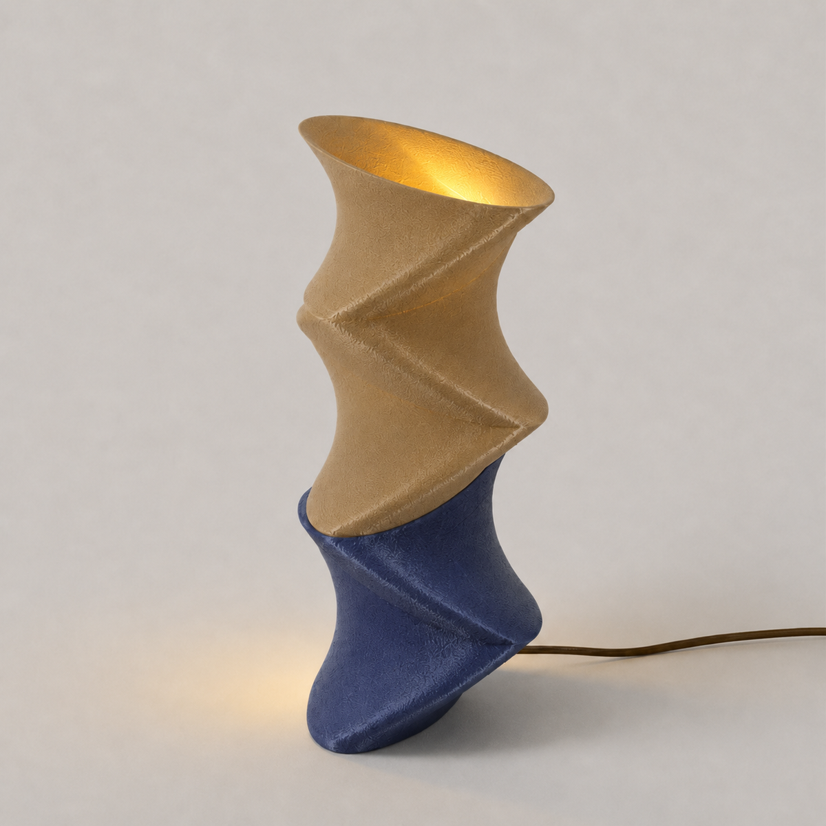 Zaha table lamp lit, showing the twisting sculptural profile in tan and deep blue with a brass cable