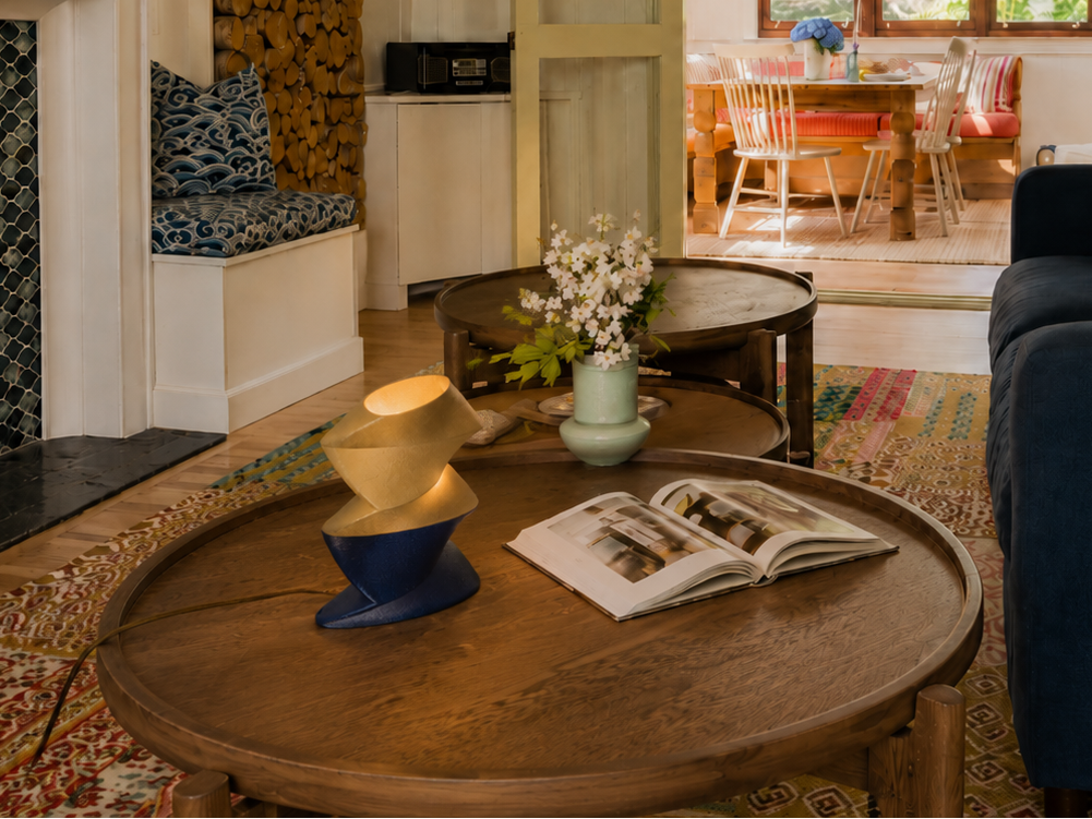 Zaha table lamp glowing on a round wooden coffee table in a living room, beside an open book and white blossoms