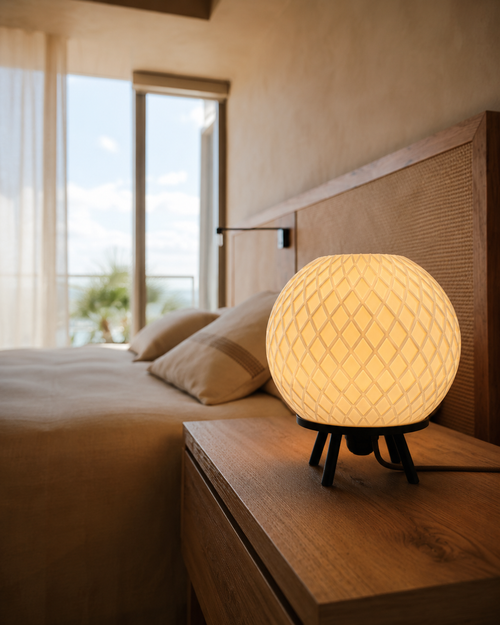 Quasar bedside lamp glowing on a wooden bedside table beside a bed with a cane headboard and a sea view behind